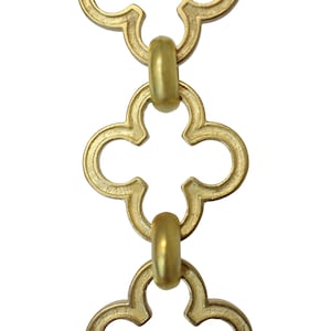 May include: Close-up of a gold-colored chain with a quatrefoil design. The chain links are connected by rounded, cylindrical connectors. The background is plain white, highlighting the chain's intricate details and metallic finish.
