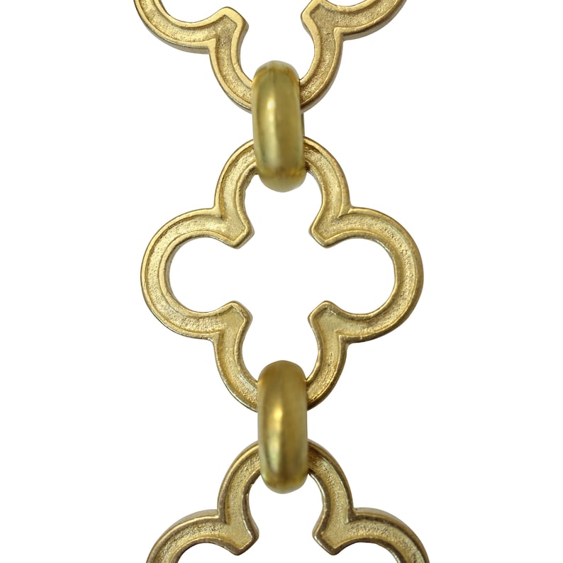 May include: Close-up of a gold-colored chain with a quatrefoil design. The chain links are connected by rounded, cylindrical connectors. The background is plain white, highlighting the chain's intricate details and metallic finish.
