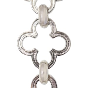 May include: Close-up of a silver-colored chain with a repeating quatrefoil design. The chain links are connected by oval-shaped connectors. The background is plain white, highlighting the intricate details of the jewelry.