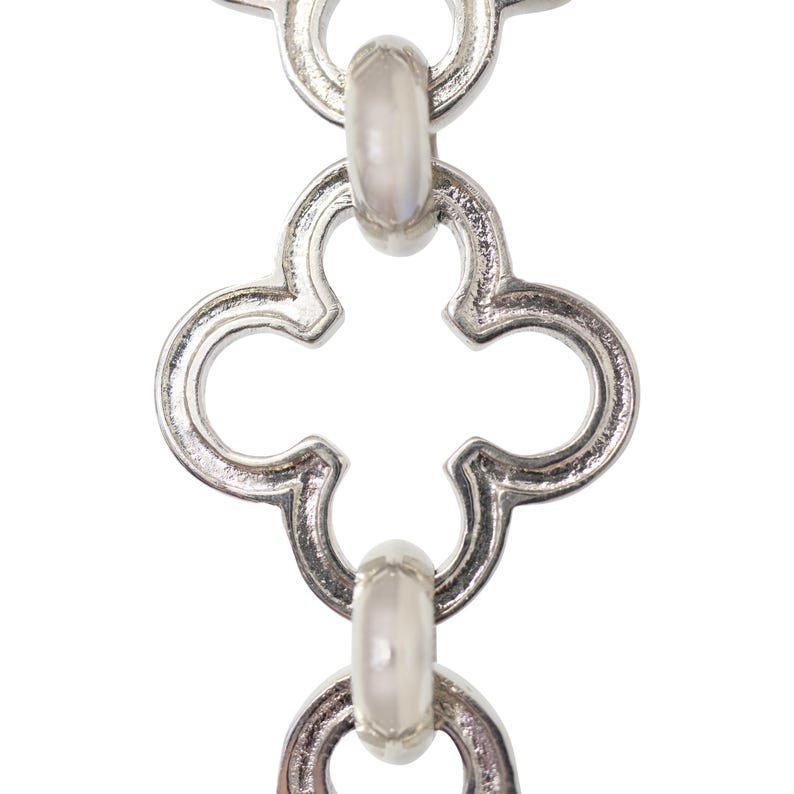 May include: Close-up of a silver-colored chain with a repeating quatrefoil design. The chain links are connected by oval-shaped connectors. The background is plain white, highlighting the intricate details of the jewelry.