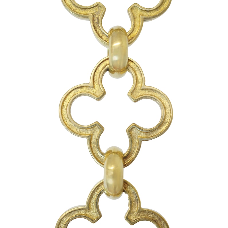 May include: Close-up of a gold-colored chain with a repeating quatrefoil design. The chain links are connected by small, rounded connectors. The background is plain white, highlighting the intricate details of the metalwork.