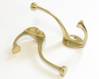 May include: Two brass-colored wall hooks. Each hook has a curved design with a rounded end and a circular base with screw holes. The hooks are made of a shiny, metallic material and are set against a white background.
