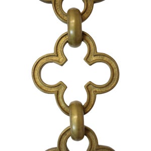 May include: Close-up of a decorative brass chain with a repeating quatrefoil design. The chain links are connected by rounded rings. The metal has a warm, antique gold finish, and the background is plain white.