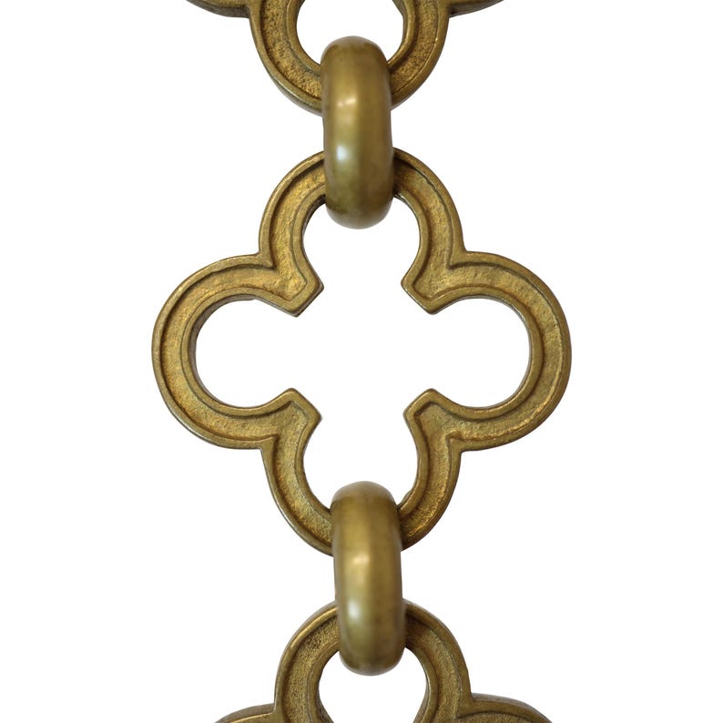 May include: Close-up of a decorative brass chain with a repeating quatrefoil design. The chain links are connected by rounded rings. The metal has a warm, antique gold finish, and the background is plain white.