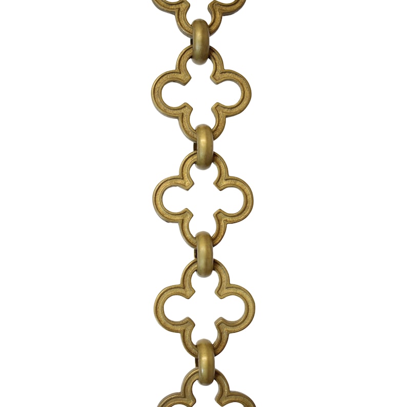 May include: Close-up of a decorative chain with a repeating quatrefoil design. The chain links are a warm, antique gold color, and the background is plain white. The chain is vertically oriented.