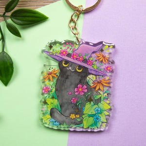 Può includere: Un portachiavi stravagante con un gatto nero che indossa un cappello da strega viola adornato di fiori. Il gatto è circondato da fiori e verde colorati. Il portachiavi ha un anello color oro.
