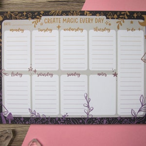 May include: A black and white weekly planner with a purple and gold floral design. The planner has a section for each day of the week, as well as a to-do list and notes section. The text "CREATE MAGIC EVERY DAY" is at the top of the planner.