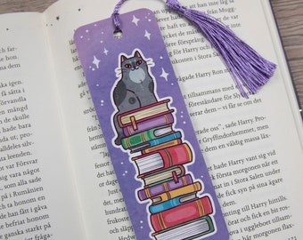 May include: Purple bookmark with a gray cat sitting on a stack of colorful books. The bookmark has a purple tassel.