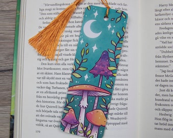May include: A teal blue bookmark with a crescent moon, stars, and three colorful mushrooms. The mushrooms have white stems and purple, orange, and pink caps.