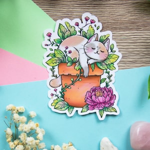May include: A sticker featuring a cartoon cat napping in a terracotta pot filled with green leaves and small pink flowers. A large pink peony is at the base of the pot. The cat is white and tan with its eyes closed.