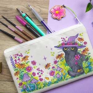 Puede incluir: Estuche de lona blanco con cremallera, con una ilustración caprichosa de un gato negro con un sombrero de bruja morado adornado con flores. El estuche está lleno de bolígrafos y lápices.