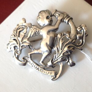 May include: Silver tone brooch featuring a cherub holding a pitcher of water, with the word "Aquarius" inscribed on a banner below. The cherub is surrounded by stylized floral designs.