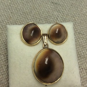 May include: A set of gold-toned earrings and a pendant featuring brown and white shell-like inlays. The earrings are round and the pendant is oval.