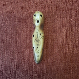 May include: A carved, elongated figurine with a rounded top and a tapered base. The beige-coloured object features several small, dark holes and etched markings. The figurine is displayed against a reddish-brown background.