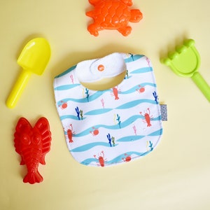 May include: A white bib with a blue and white wave pattern and red lobsters. The bib has a white snap closure. There are also four plastic toys: a yellow shovel, a green rake, a red lobster, and an orange turtle.