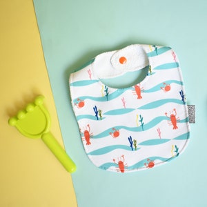 May include: Two white baby bibs with different patterns. One bib has a blue and white pattern with lobsters and seaweed. The other bib has a white background with a small, colorful floral pattern.