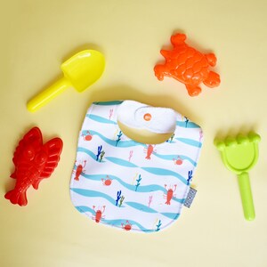 May include: A white bib with a blue and white wave pattern and red lobsters. The bib has a white snap closure. There are also four plastic toys: a yellow shovel, a green rake, a red lobster, and an orange turtle.