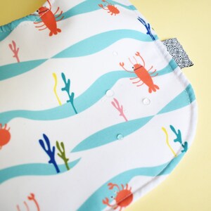 May include: A white bib with a blue and white wavy pattern. The bib features a red lobster, a crab, and seaweed in a sea-themed design.