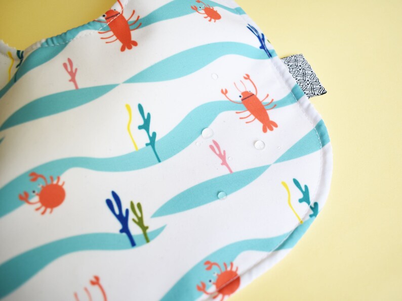May include: A white bib with a blue and white wavy pattern. The bib features a red lobster, a crab, and seaweed in a sea-themed design.