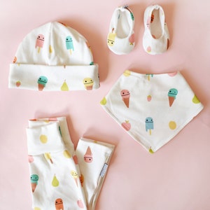 May include: A white baby hat, pants, and bib with a pattern of ice cream cones, popsicles, pears, and strawberries. The set also includes a pair of white baby shoes with a similar pattern.