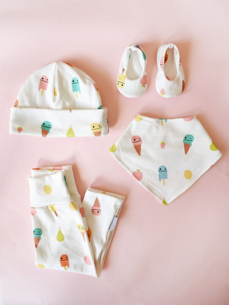 May include: A white baby hat, pants, and bib with a pattern of ice cream cones, popsicles, pears, and strawberries. The set also includes a pair of white baby shoes with a similar pattern.
