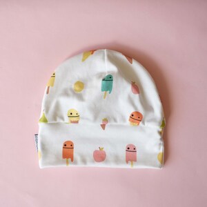 May include: White cotton beanie with a colorful pattern of ice cream cones and fruit. The ice cream cones have smiling faces.