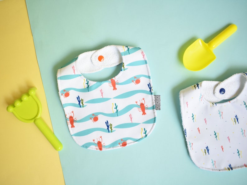 May include: Two white baby bibs with different patterns. One bib has a blue and white pattern with lobsters and seaweed. The other bib has a white background with a small, colorful floral pattern.