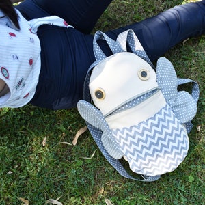 May include: A white and gray fabric backpack shaped like a fish with large button eyes. The backpack has a zippered opening and a chevron pattern on the bottom.