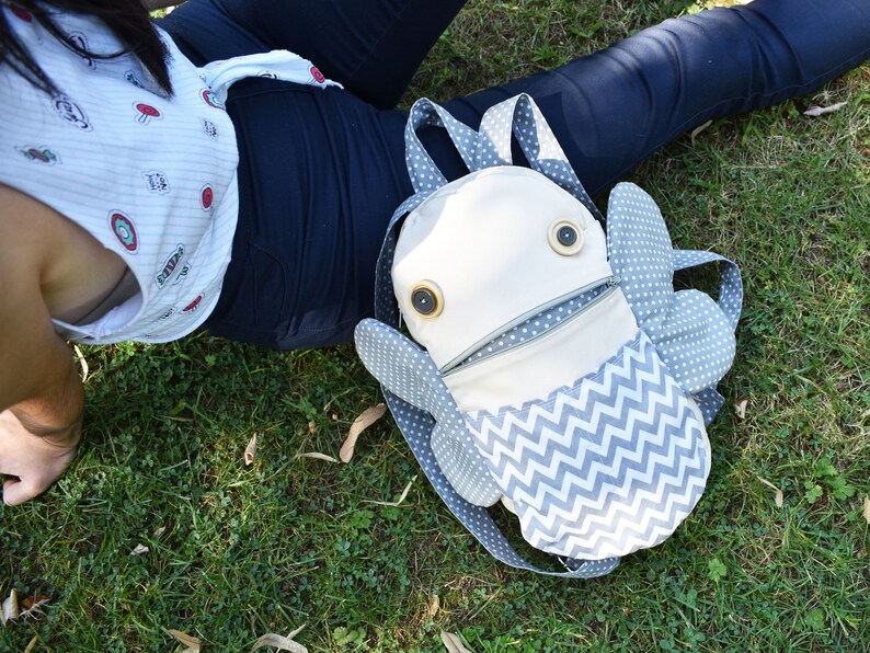 May include: A white and gray fabric backpack shaped like a fish with large button eyes. The backpack has a zippered opening and a chevron pattern on the bottom.