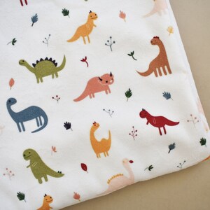 May include: White blanket with a colorful dinosaur pattern. The blanket features various dinosaurs in shades of orange, green, pink, and blue, along with small leaf and sprig accents. The blanket is folded, showing a corner.