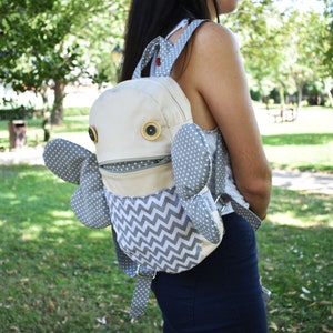 May include: A beige and gray backpack with a whimsical design. The backpack has a large, round, white face with two large, brown eyes and a zippered mouth. The backpack has two gray wings with white polka dots. The backpack is worn by a person in a park.