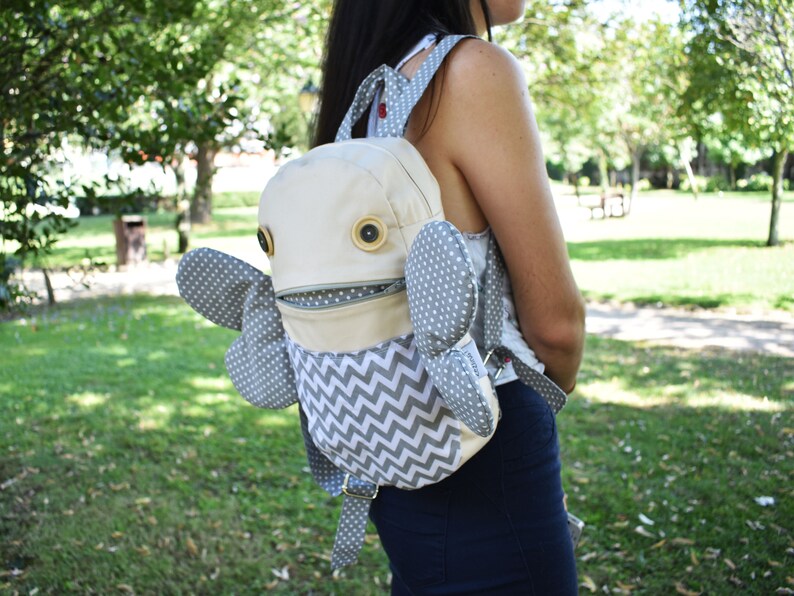 May include: A beige and gray backpack with a whimsical design. The backpack has a large, round, white face with two large, brown eyes and a zippered mouth. The backpack has two gray wings with white polka dots. The backpack is worn by a person in a park.