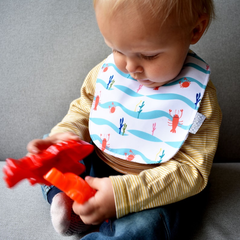 May include: A baby wearing a white bib with a blue and white wave pattern and red lobsters and crabs. The bib has a silver glitter trim. The baby is holding a red plastic toy.