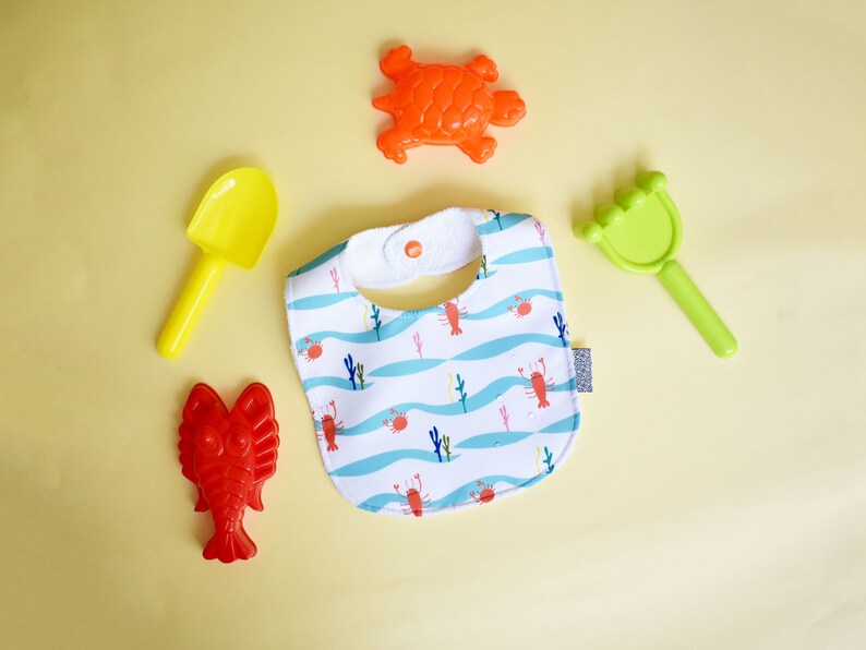 May include: A white bib with a blue and white wave pattern and red lobsters. The bib has a white snap closure. There are also four plastic toys: a yellow shovel, a green rake, a red lobster, and an orange turtle.