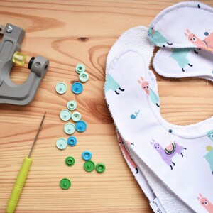May include: A set of two white baby bibs with a llama print. The bibs are made of a soft, absorbent material and have a terry cloth backing. The bibs are shown with a snap press, a tool used to attach snaps to fabric, and a variety of colorful snaps.