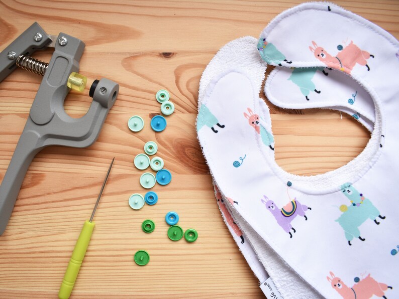 May include: A set of two white baby bibs with a llama print. The bibs are made of a soft, absorbent material and have a terry cloth backing. The bibs are shown with a snap press, a tool used to attach snaps to fabric, and a variety of colorful snaps.
