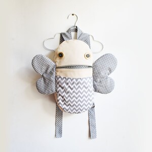 May include: A gray and white patterned backpack with a zippered pocket and two large wings. The backpack has a whimsical, cartoon-like face with two large black eyes and a zipper for a mouth.