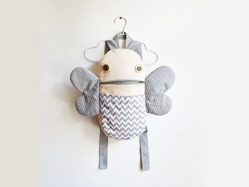 May include: A gray and white patterned backpack with a zippered pocket and two large wings. The backpack has a whimsical, cartoon-like face with two large black eyes and a zipper for a mouth.