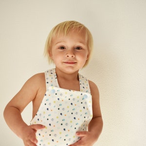 May include: A young child with blonde hair wearing a white overall with a blue, green, and orange pattern. The overall has two blue buttons on the straps.