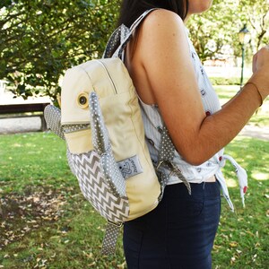 May include: A light beige backpack with a gray and white chevron pattern. The backpack has a large, round, white button on the front and two gray, fabric ears. The backpack has a small, white tag with the word "Zest" on it.