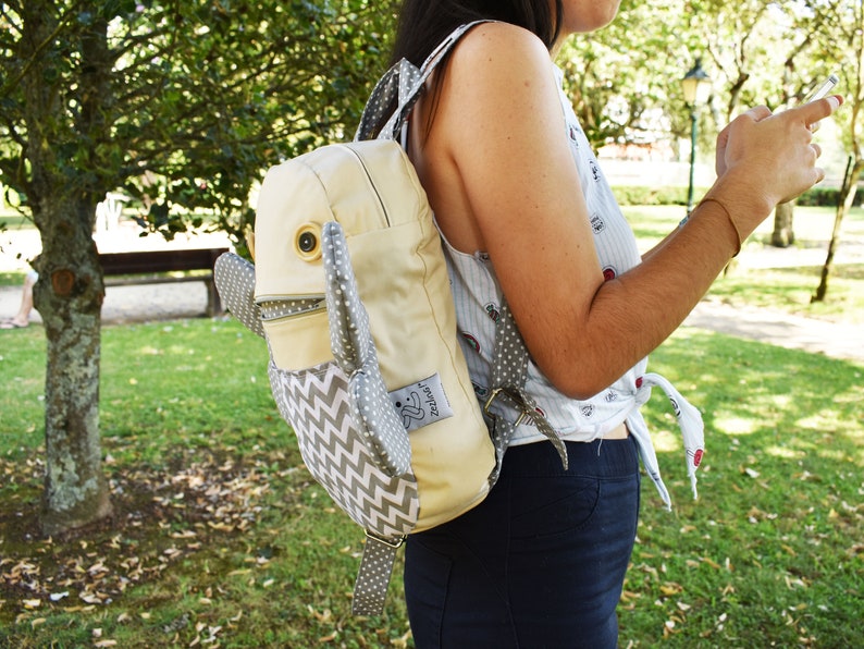 May include: A light beige backpack with a gray and white chevron pattern. The backpack has a large, round, white button on the front and two gray, fabric ears. The backpack has a small, white tag with the word "Zest" on it.