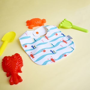 May include: A white bib with a blue and white wave pattern and red lobsters. The bib has a white snap closure. There are also red and orange plastic toys in the image.
