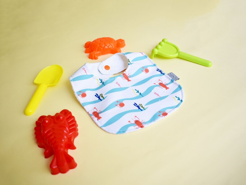 May include: A white bib with a blue and white wave pattern and red lobsters. The bib has a white snap closure. There are also red and orange plastic toys in the image.