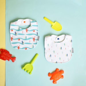 May include: Two white cotton baby bibs with different patterns. One bib has a blue and white wave pattern with red crabs. The other bib has a white background with a repeating pattern of colorful cacti.  The bibs are on a light blue background with a yellow shovel, a red fish, a green rake, and an orange turtle.