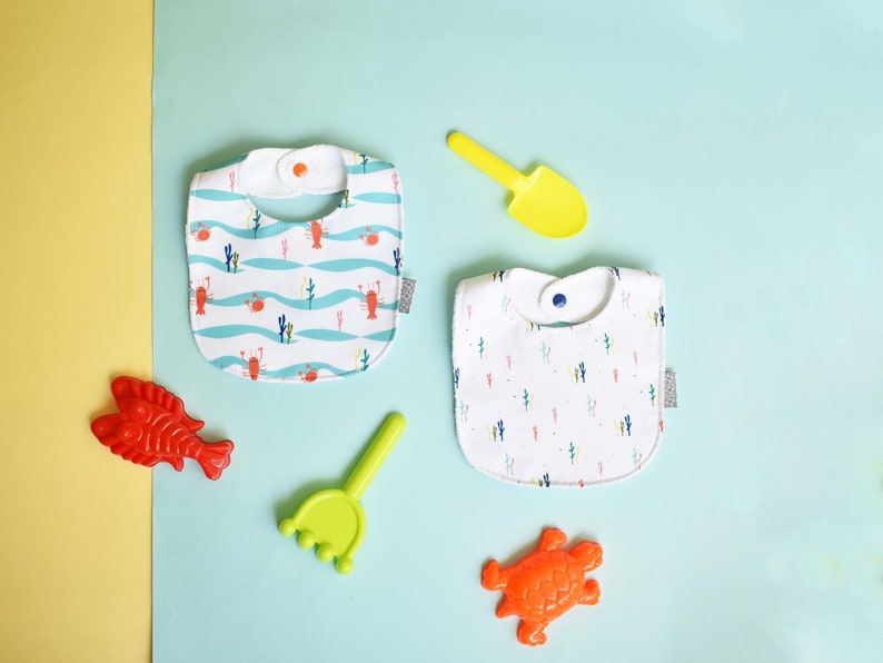 May include: Two white cotton baby bibs with different patterns. One bib has a blue and white wave pattern with red crabs. The other bib has a white background with a repeating pattern of colorful cacti.  The bibs are on a light blue background with a yellow shovel, a red fish, a green rake, and an orange turtle.