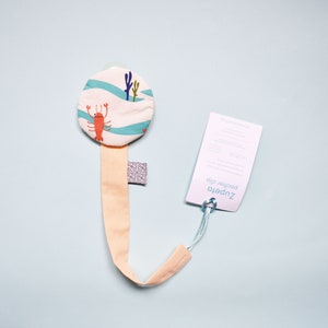 May include: A light pink dummy clip with a round, white fabric attachment featuring a blue and white wave pattern and a red lobster design. The clip is attached to a white tag with the text "Zupetto Pacifier Clip".