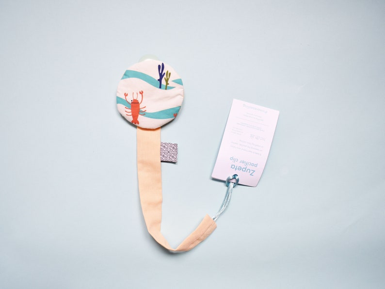 May include: A light pink dummy clip with a round, white fabric attachment featuring a blue and white wave pattern and a red lobster design. The clip is attached to a white tag with the text "Zupetto Pacifier Clip".
