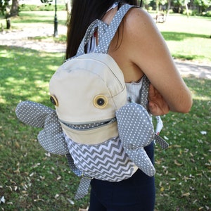 May include: A gray and white patterned backpack with large, round, brown eyes and a zippered mouth. The backpack has gray polka dot wings.