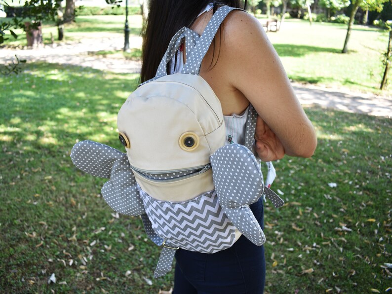 May include: A gray and white patterned backpack with large, round, brown eyes and a zippered mouth. The backpack has gray polka dot wings.