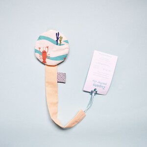 May include: A light pink dummy clip with a round, white fabric attachment featuring a blue and white wave pattern and a red lobster design. The clip is attached to a white tag with the text "Zupetto Pacifier Clip".